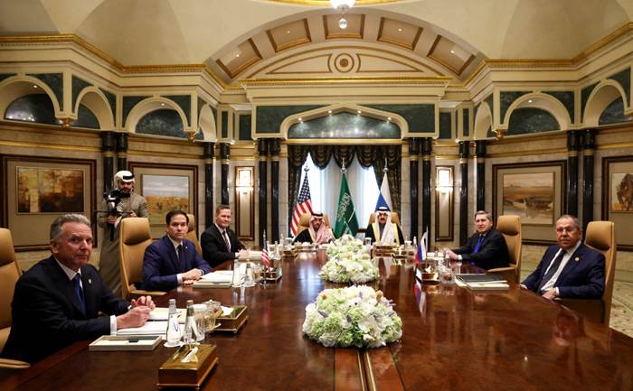 Men sitting around a table with multiple floral centerpieces in a richly decorated room.
